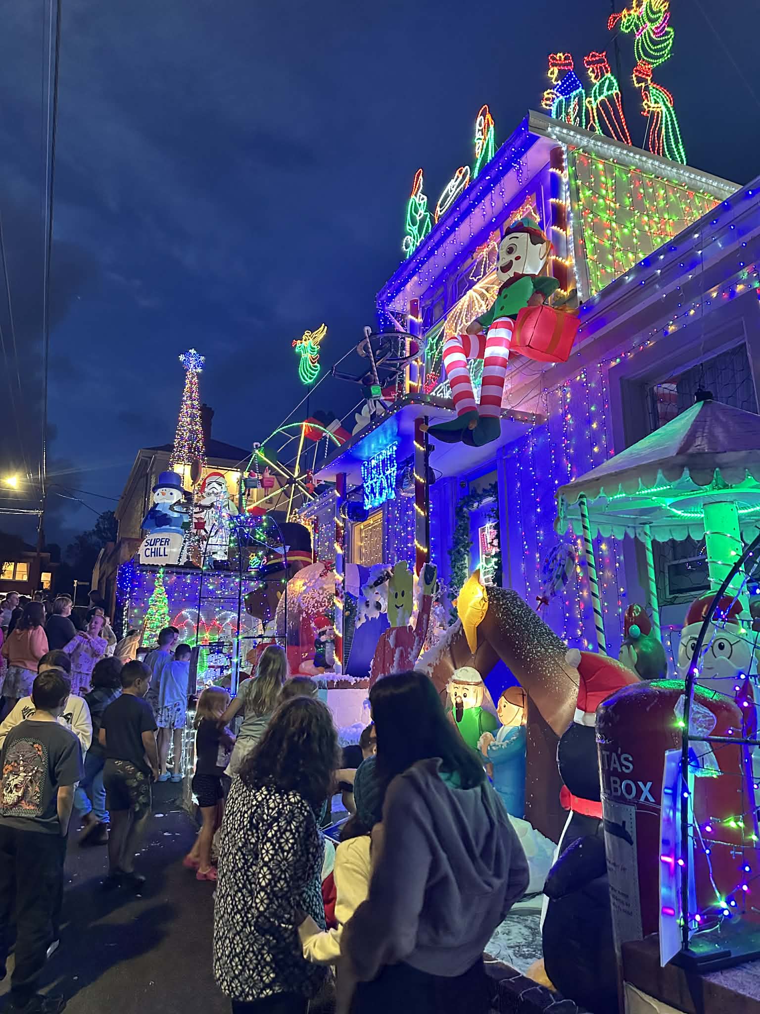 Johnnys Christmas Lights Display filled with people viewing a house decorated with bright Christmas lights and holiday ornaments. The building features large illuminated figures, colorful string lights, and festive displays against a dark blue evening sky in Hobart, Tasmania.