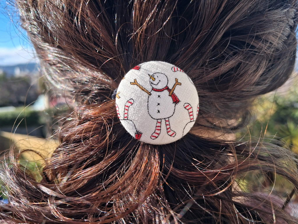 A round fabric christmas hair tie with a snowman design, shown pinned to a brown hair bun outdoors.